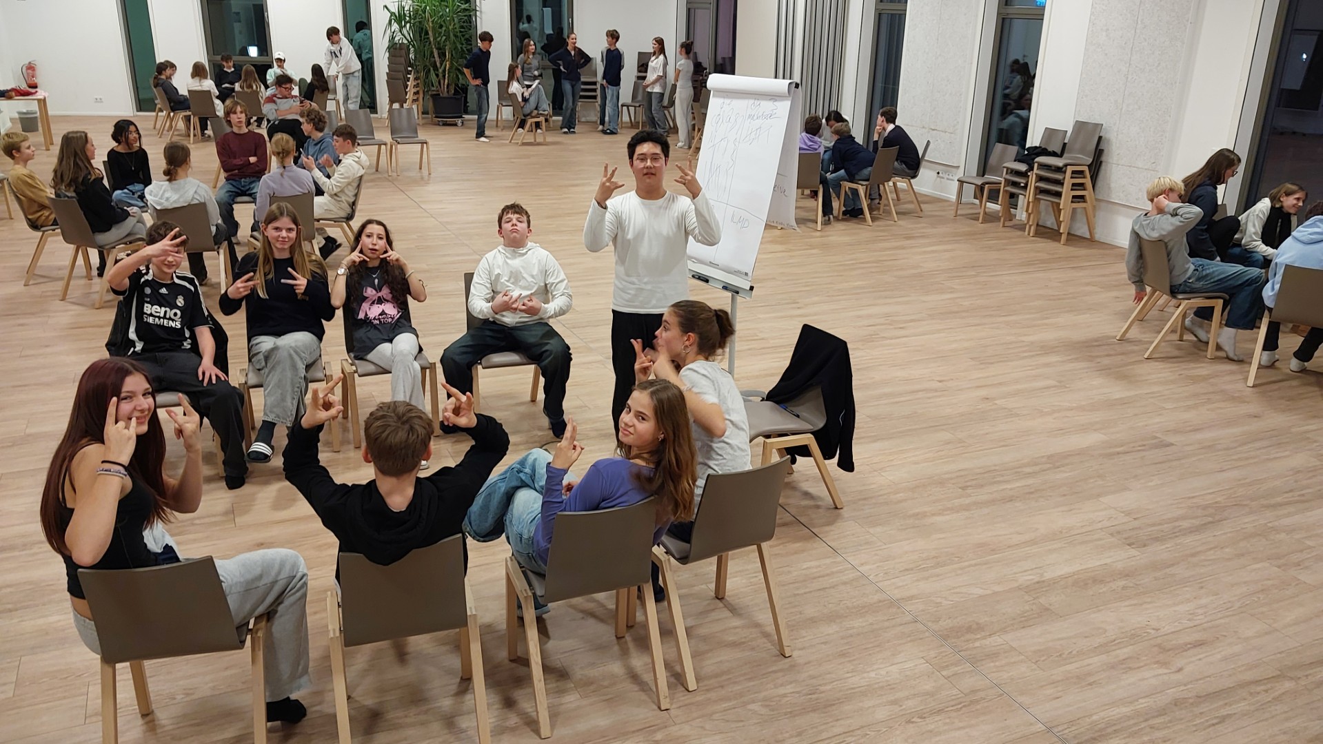 Vielen Jugendliche in einem Saal. Im Vordergrund eine kleine Gruppe, die vor einem Flipchart sitzen und offenbar Freude an der Arbeit haben.
